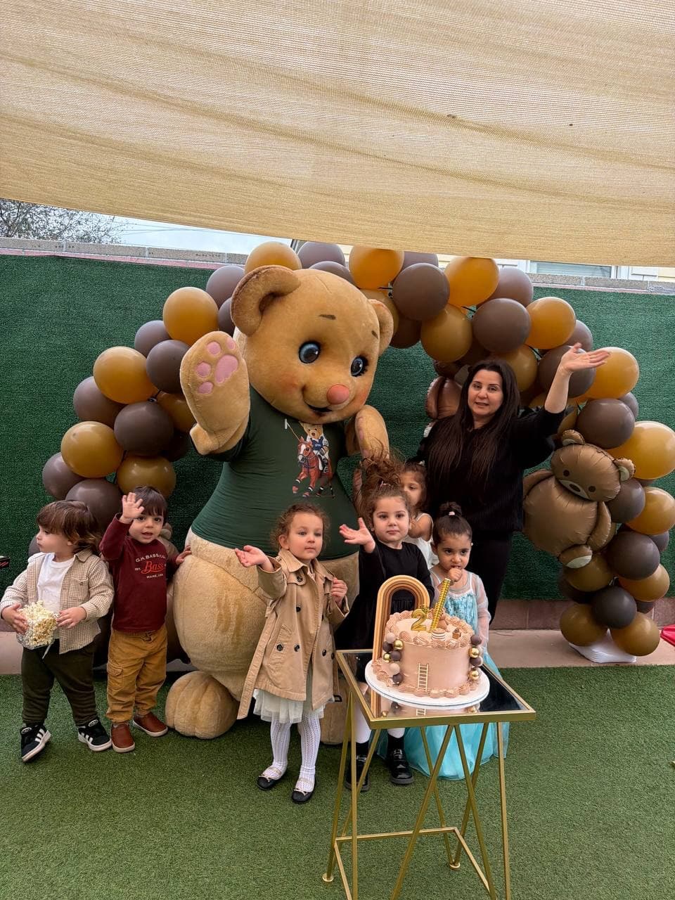 Children celebrating a birthday with a teddy bear mascot, balloons, and cake