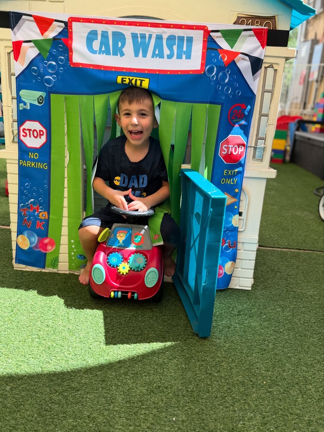 Toddler on a toy car in front of a playful Car Wash playhouse